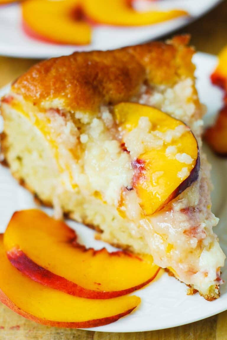 A slice of peach cake topped with peach slices and crumbly topping, served on a white plate with additional fresh peach slices on the side.