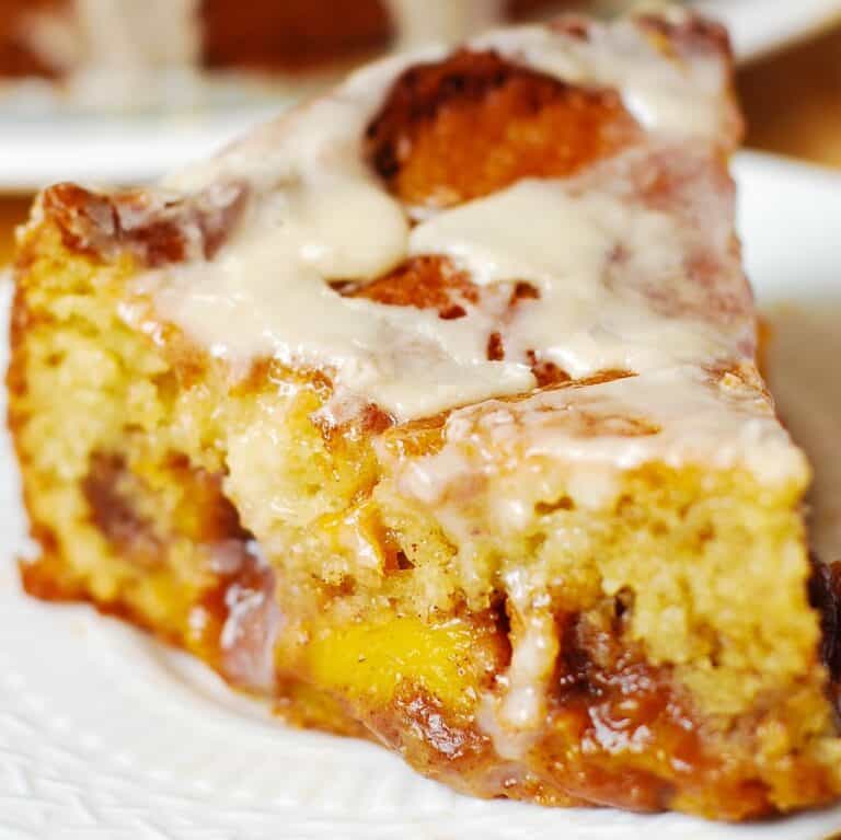 A close-up of a slice of peach coffee cake with chunks of peaches, topped with a drizzle of creamy glaze, served on a white plate.