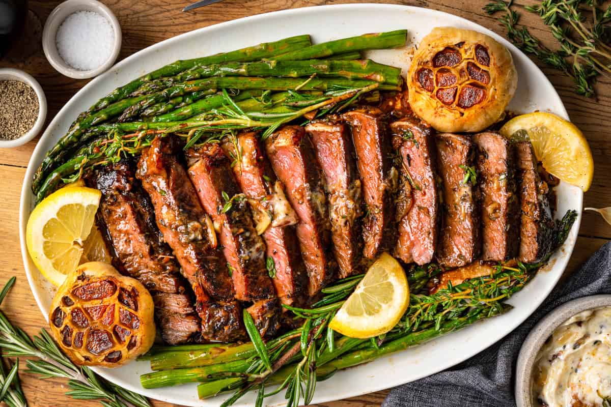 15 Summer Grilling Recipes So Good You'll Make Them Twice A platter of sliced grilled steak topped with herbs and butter, surrounded by roasted garlic, lemon slices, and asparagus spears, garnished with fresh rosemary and thyme.