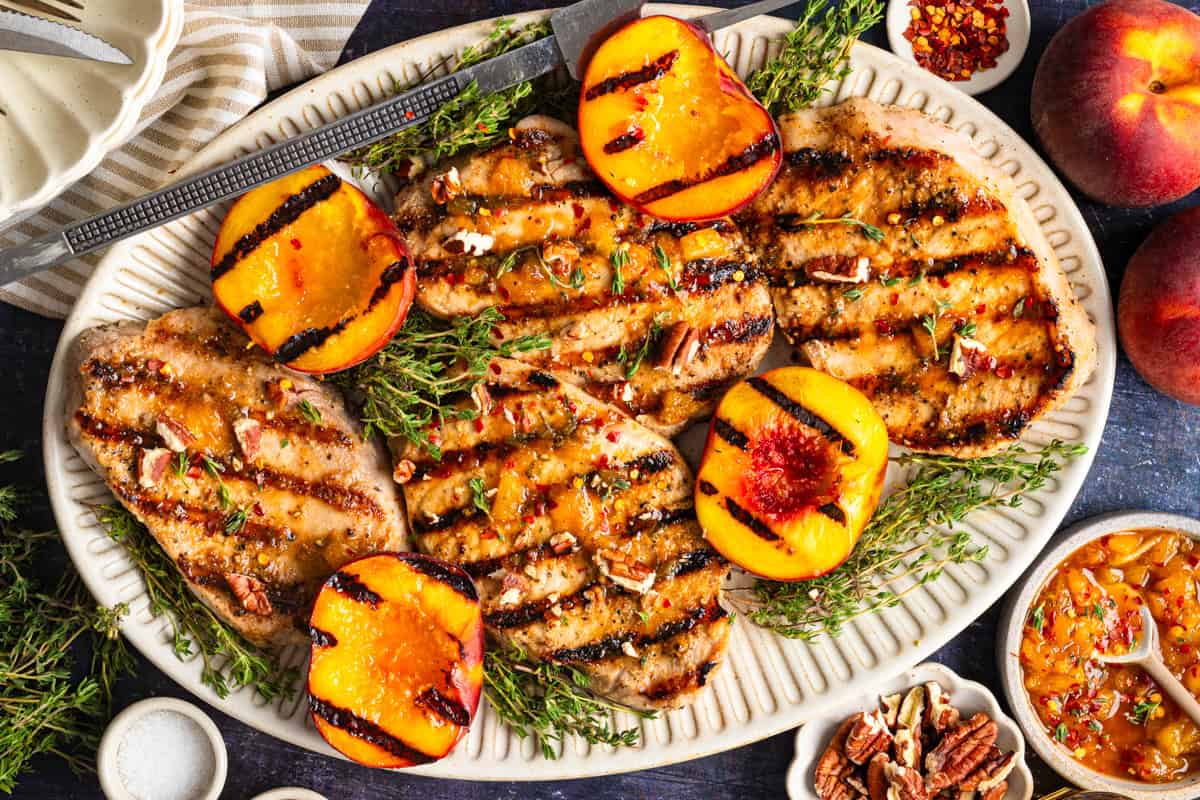 15 Summer Grilling Recipes So Good You'll Make Them Twice A platter with grilled pork chops and halved grilled peaches, garnished with fresh herbs. Small bowls of chopped pecans, red pepper flakes, and chutney surround the platter on a dark tabletop.