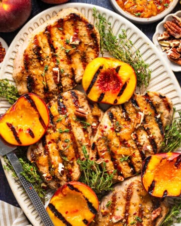 A platter of grilled pork chops garnished with fresh herbs, surrounded by grilled peach halves, chopped pecans, and a small bowl of sauce, set on a dark table.