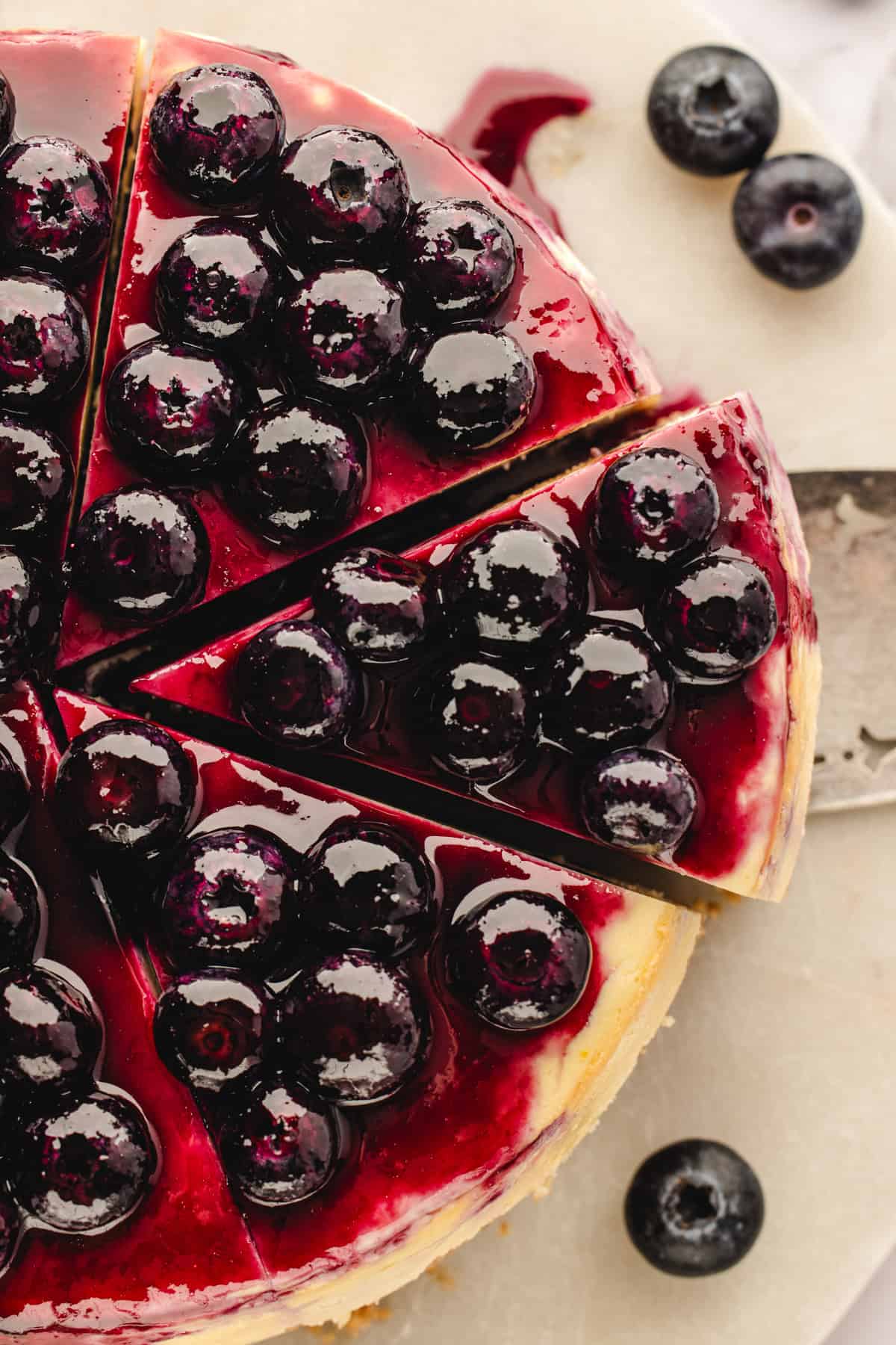 Top-down view of a blueberry cheesecake with a glossy blueberry topping, sliced with one piece partially removed, and a few loose blueberries on a light-colored surface.