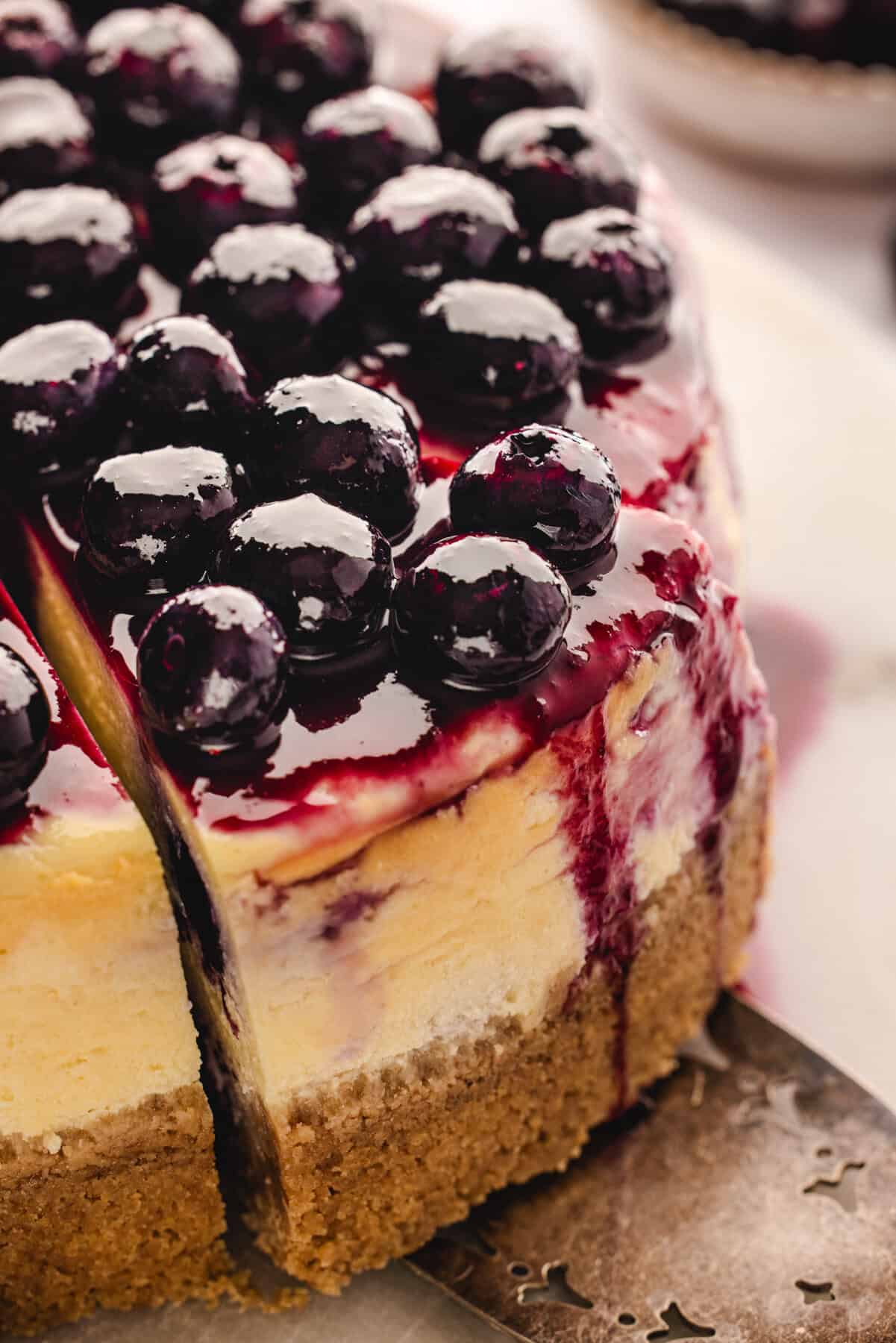 A close-up of a cheesecake topped with glossy blueberries and blueberry sauce, with a slice being lifted out, showing the creamy layers and graham cracker crust.
