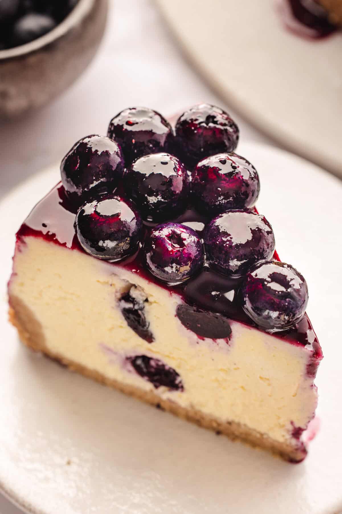 A slice of creamy cheesecake topped with glossy, fresh blueberries and a layer of blueberry glaze, sitting on a white plate.