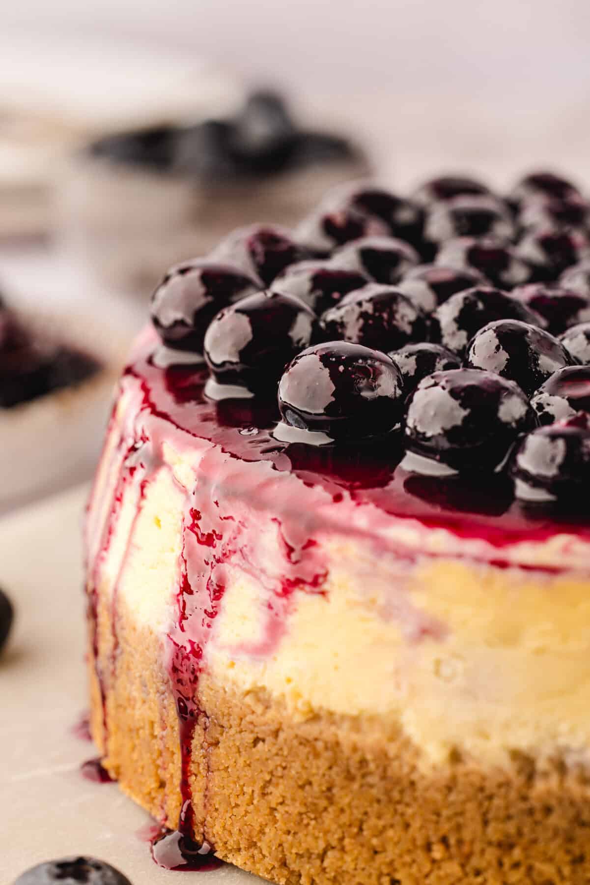 A close-up of a cheesecake topped with glossy blueberries and blueberry sauce, with sauce dripping down the side of the creamy filling and graham cracker crust.