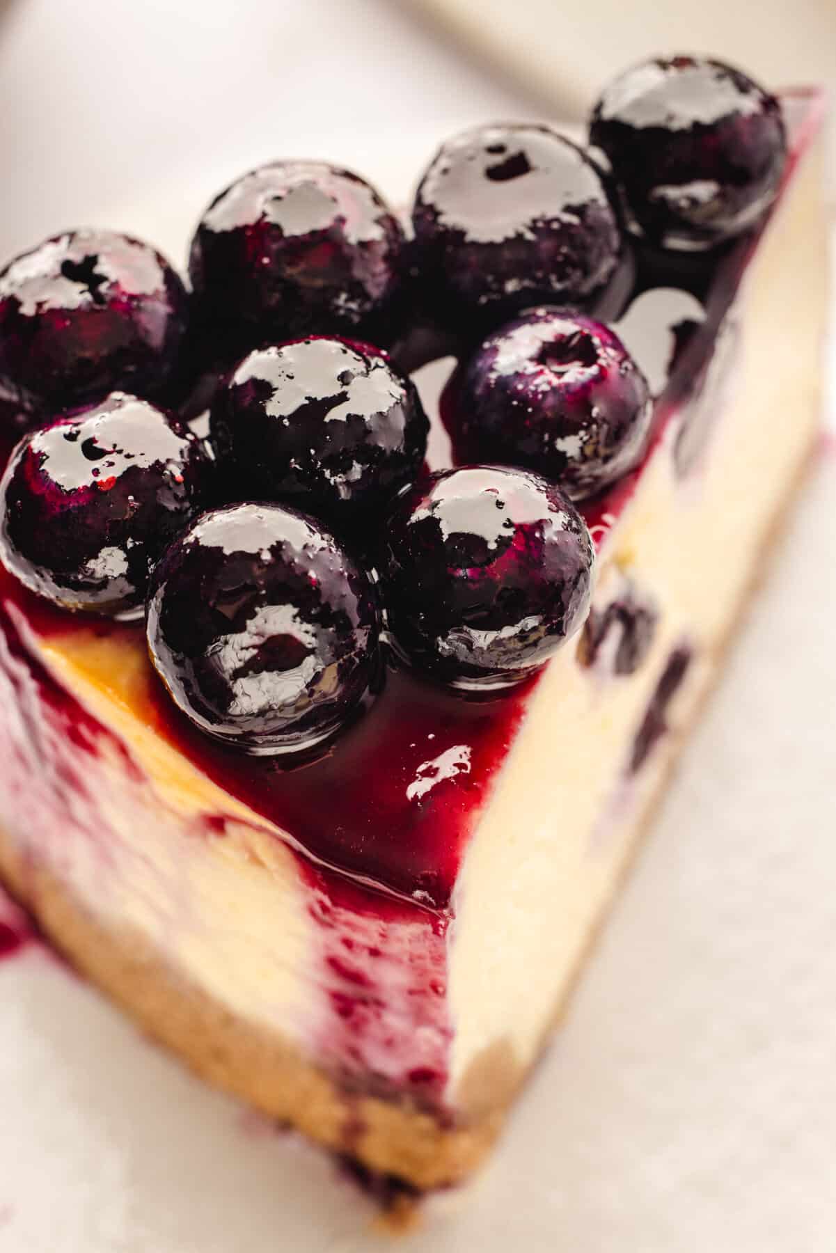 A close-up of a slice of cheesecake topped with glossy, fresh blueberries and blueberry sauce, showing the creamy texture and a crumbly graham cracker crust.