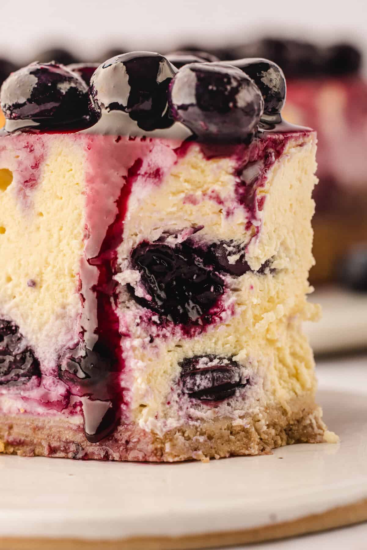 A close-up of a thick slice of cheesecake topped with glossy blueberries and blueberry sauce, with visible whole blueberries inside the creamy filling and a crumbly graham cracker crust at the bottom.
