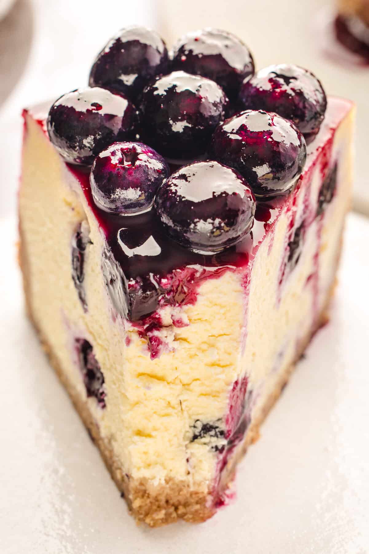 A slice of creamy cheesecake topped with glossy blueberry sauce and whole blueberries, with a graham cracker crust visible at the base.