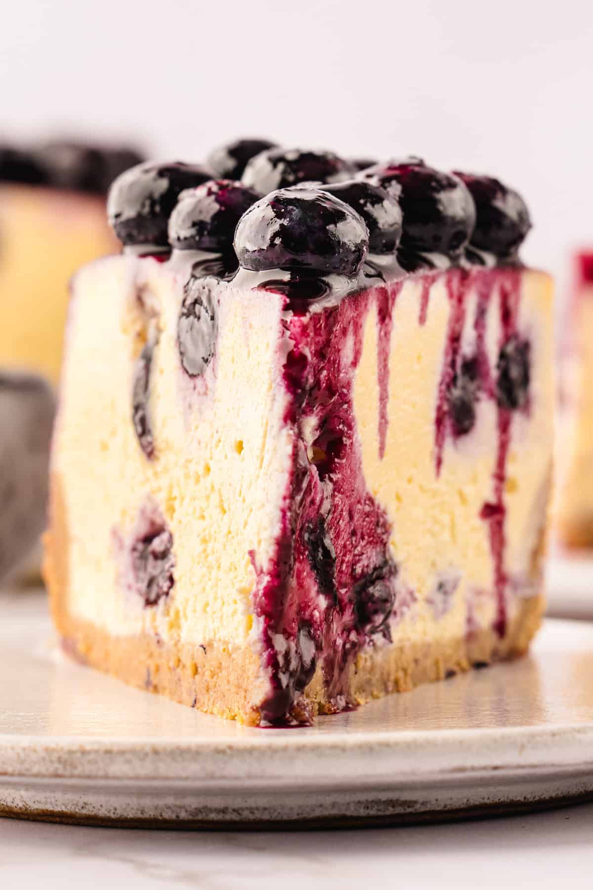 A close-up of a slice of blueberry cheesecake with a graham cracker crust, creamy filling, and blueberry topping, with blueberry sauce dripping down the sides, served on a white plate.