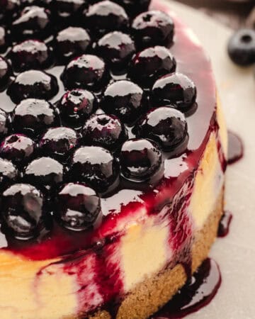 A close-up of a cheesecake topped with glossy, fresh blueberries and blueberry sauce, with sauce dripping down the sides onto a light surface.