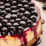 Close-up of a cheesecake topped with glossy, fresh blueberries and blueberry sauce, with the sauce dripping down the sides onto a brown crumb crust.
