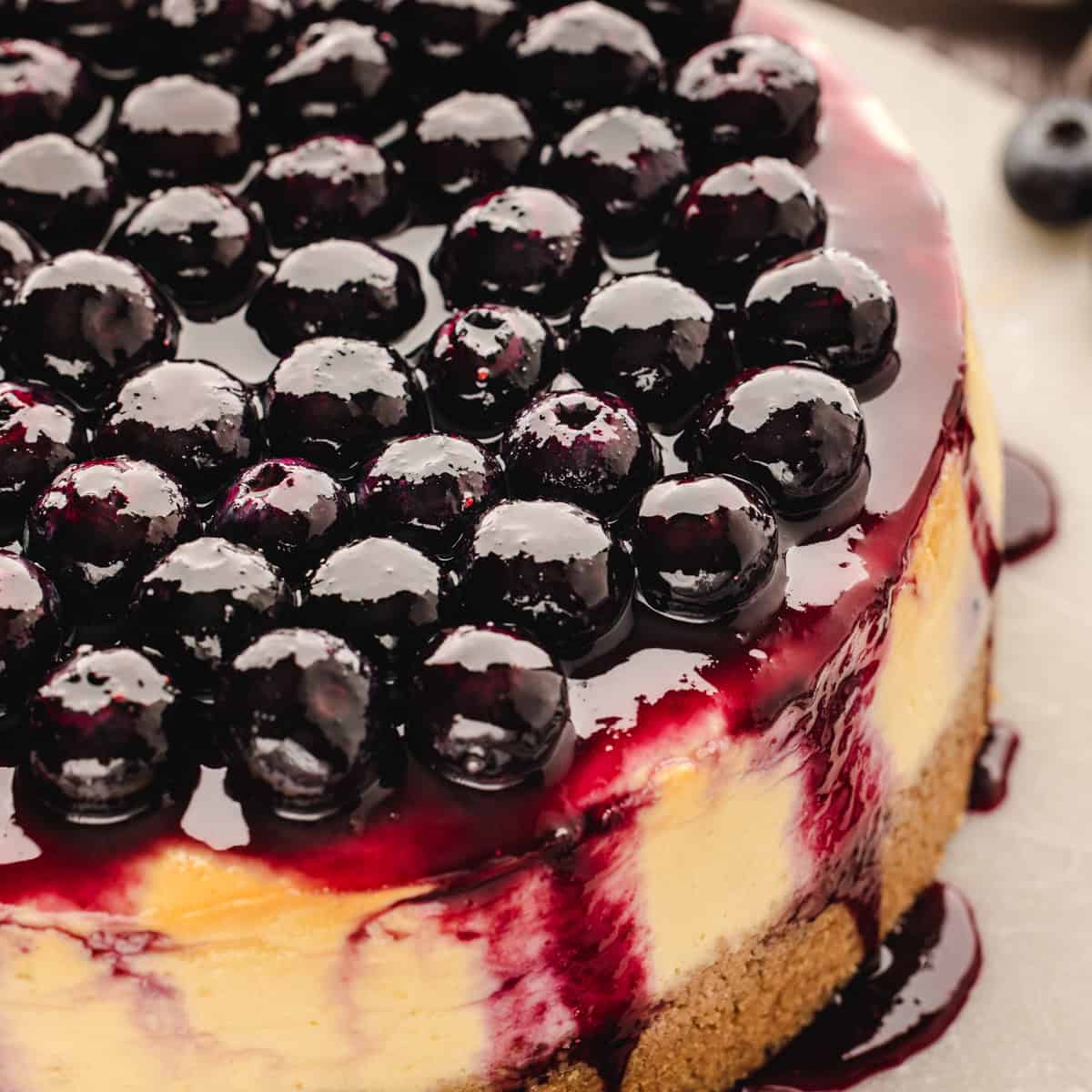 Close-up of a cheesecake topped with glossy, fresh blueberries and blueberry sauce, with the sauce dripping down the sides onto a brown crumb crust.