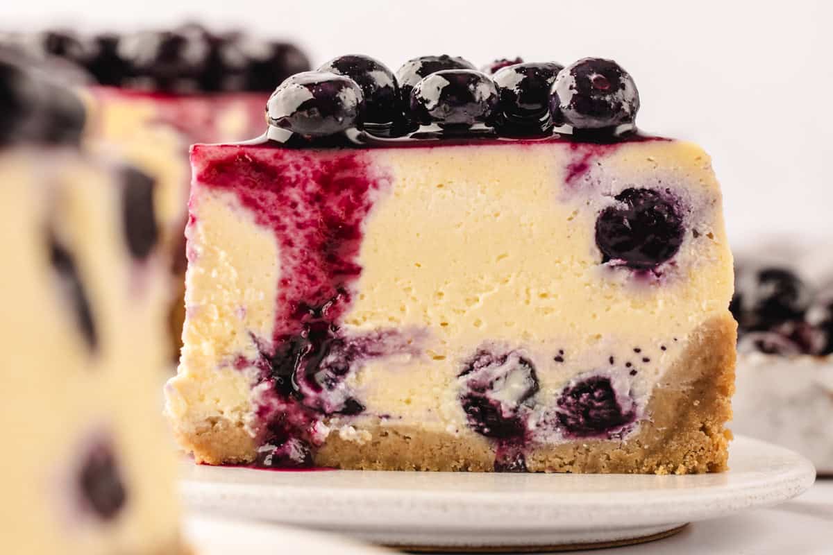 A close-up of a slice of blueberry cheesecake with a graham cracker crust, creamy filling swirled with blueberries, and topped with whole blueberries and purple sauce, set on a white plate.