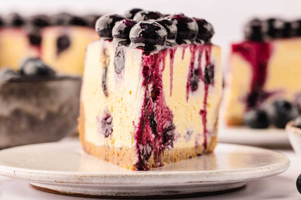 A slice of blueberry cheesecake with a golden crust, creamy filling, blueberry swirls, and fresh blueberries on top, drizzled with purple blueberry sauce, served on a white plate.
