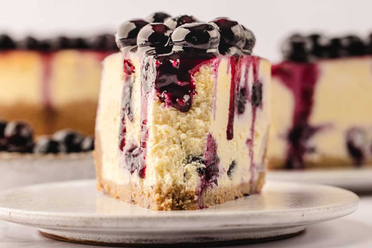 A close-up of a thick slice of cheesecake with a golden crust, topped with glossy blueberry sauce and whole blueberries, served on a white plate. The cheesecake has visible berry swirls and a creamy texture.