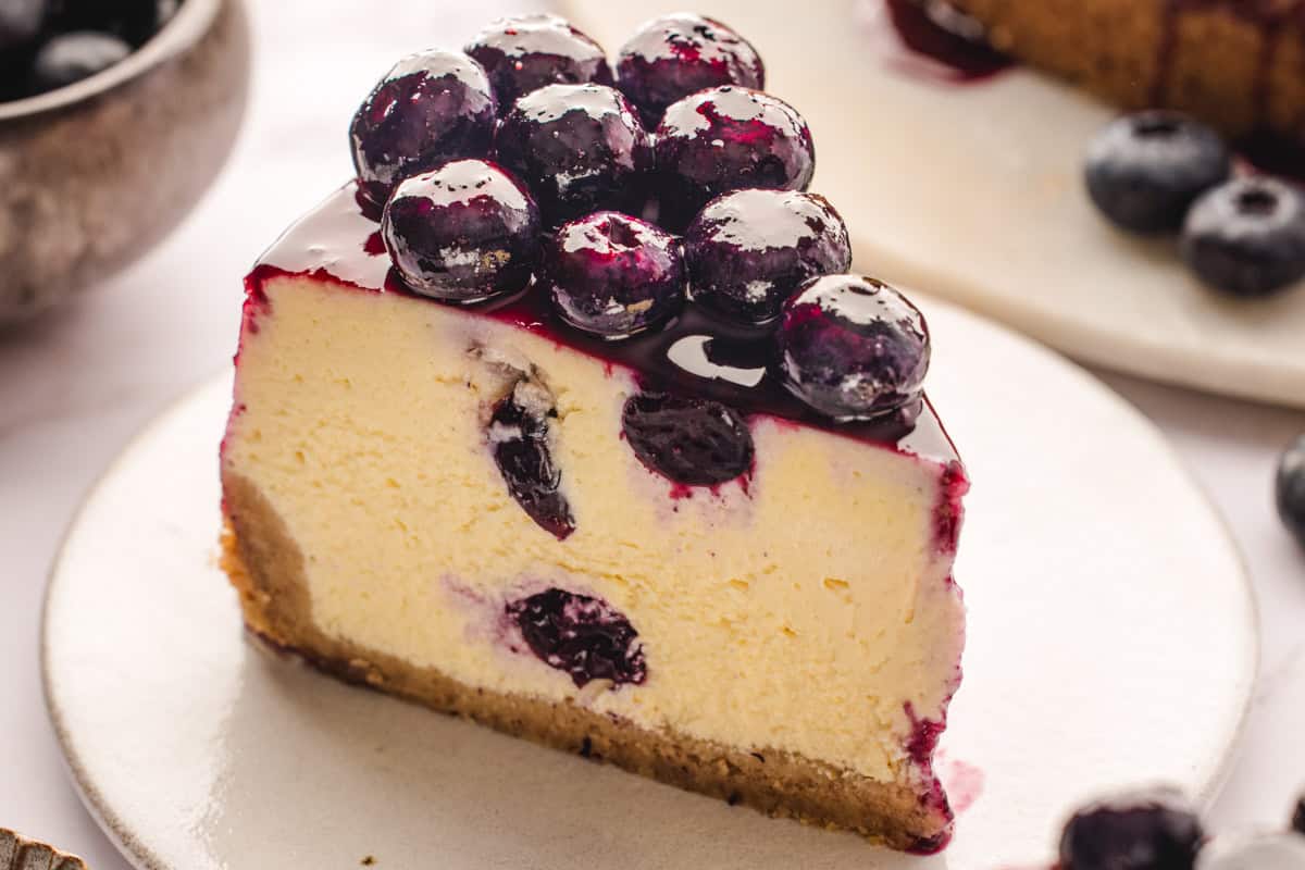 A slice of creamy cheesecake with a graham cracker crust, topped with glossy fresh blueberries and blueberry sauce, sits on a white plate. Some blueberries are baked into the cheesecake.