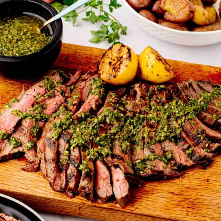 Sliced grilled steak topped with green chimichurri sauce on a wooden board, served with charred lemon halves. A bowl of potatoes and a small bowl of extra chimichurri are in the background.