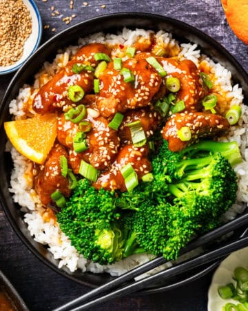 A bowl of white rice topped with orange chicken, steamed broccoli, sesame seeds, and chopped green onions, garnished with an orange slice and served with black chopsticks.