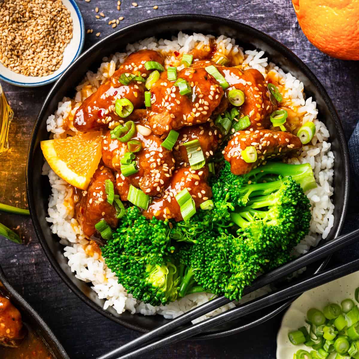 A bowl of white rice topped with orange chicken, steamed broccoli, sesame seeds, and chopped green onions, garnished with an orange slice and served with black chopsticks.