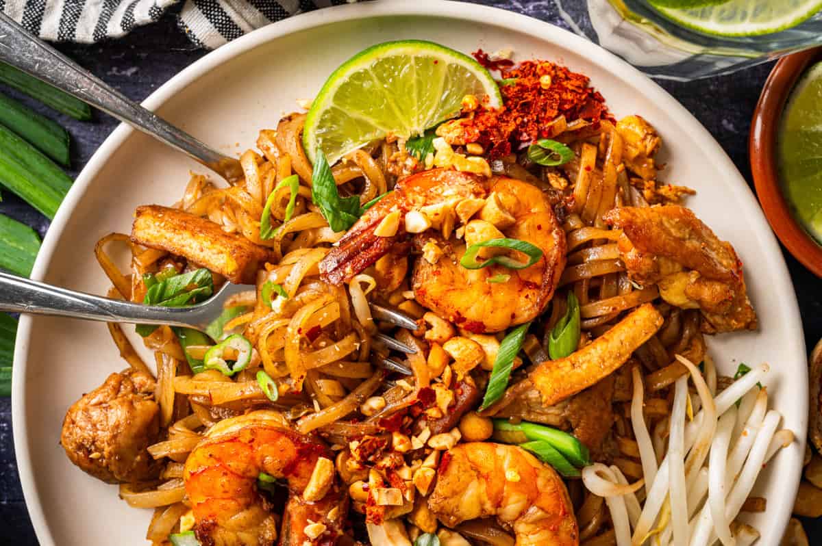 A plate of Pad Thai topped with shrimp, chicken, green onions, crushed peanuts, chili flakes, and a lime wedge. Bean sprouts are on the side, and a fork and spoon are placed in the noodles.
