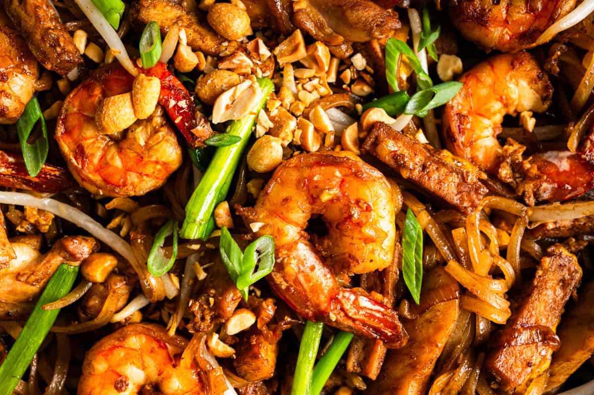 Close-up of pad Thai with shrimp, chicken, green onions, crushed peanuts, and noodles, all coated in a glossy sauce. The dish is vibrant and topped with fresh green slices and crunchy nuts.