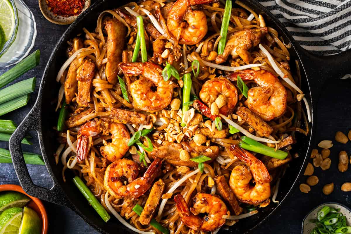 A skillet filled with shrimp and chicken Pad Thai topped with peanuts, green onions, and bean sprouts, surrounded by lime wedges and extra peanuts on a dark table.