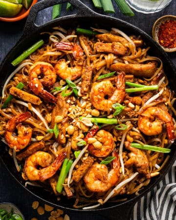 A cast iron skillet filled with shrimp and chicken pad thai, garnished with chopped peanuts, green onions, and bean sprouts, with lime wedges and chili flakes on the side.