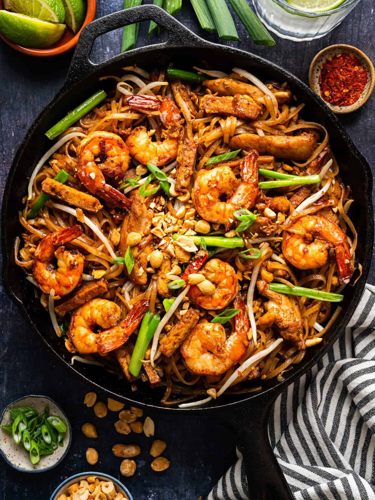 A skillet filled with shrimp and chicken pad thai, garnished with peanuts, green onions, and bean sprouts. Lime wedges, chili flakes, and extra peanuts are arranged around the skillet on a dark surface.
