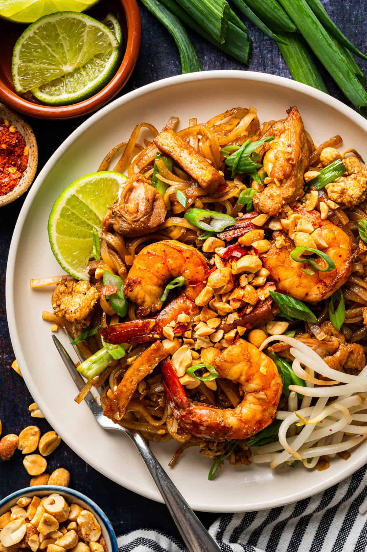 A plate of Pad Thai topped with shrimp, chicken, chopped peanuts, green onions, and bean sprouts, garnished with lime wedges. Surrounding the plate are fresh lime, peanuts, chili flakes, and scallions.