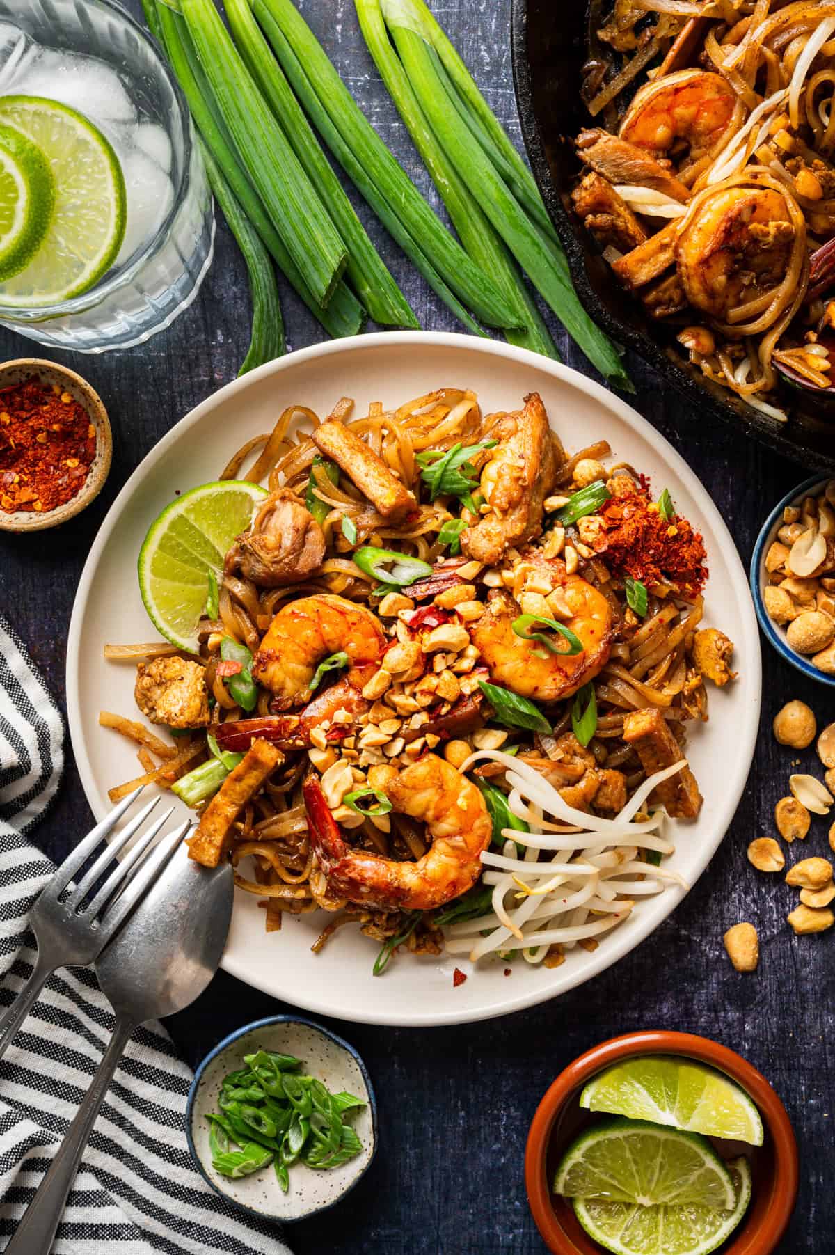 A plate of Pad Thai topped with shrimp, chicken, bean sprouts, peanuts, green onions, and lime wedges, with a side of chopped peanuts, green onions, chili flakes, and a glass of water with lime slices on the table.