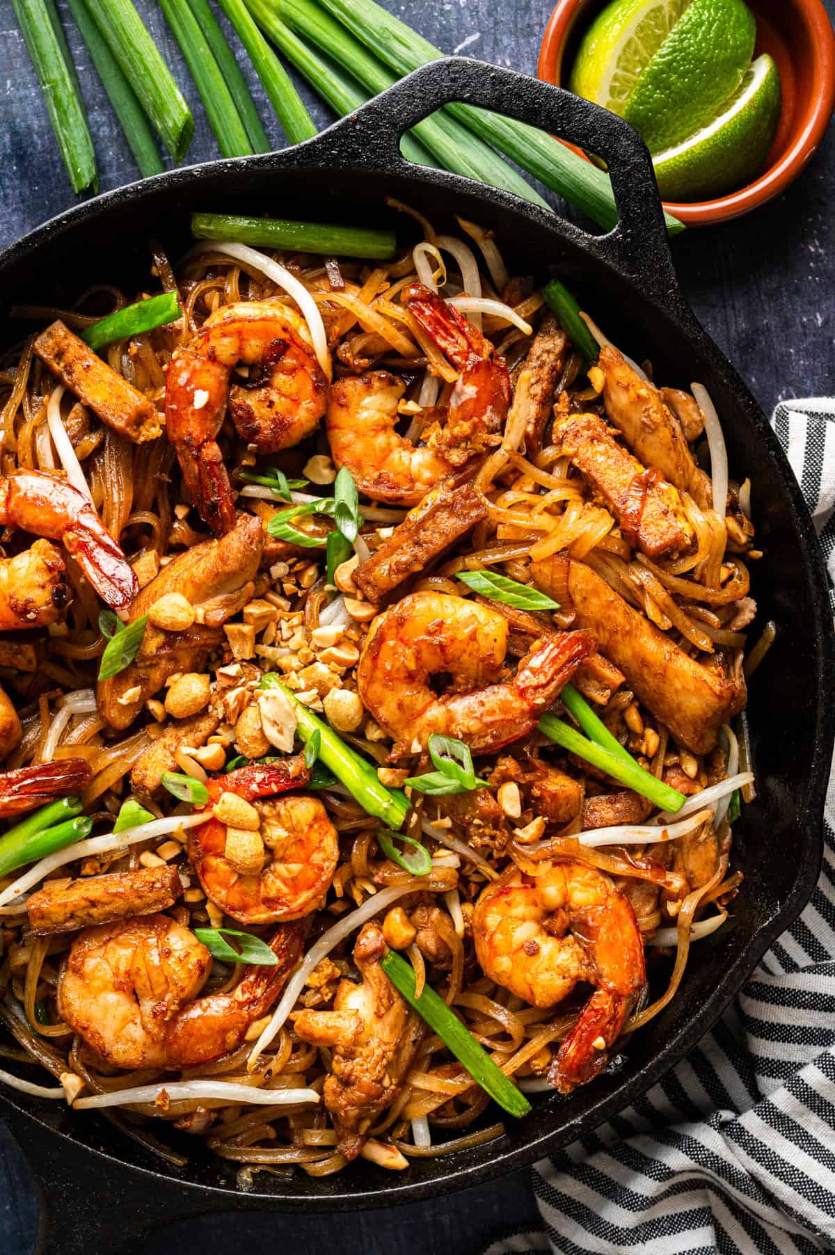 A skillet filled with pad thai topped with shrimp, chicken, tofu, bean sprouts, chopped peanuts, and green onions. Fresh lime wedges and green onions are nearby on a dark surface.
