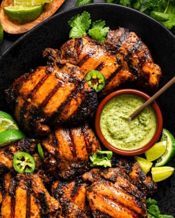Grilled Peruvian chicken thighs with char marks on a black plate, garnished with lime wedges, cilantro, and sliced jalape&ntilde;o. A small bowl of creamy green sauce sits on the plate beside the chicken.