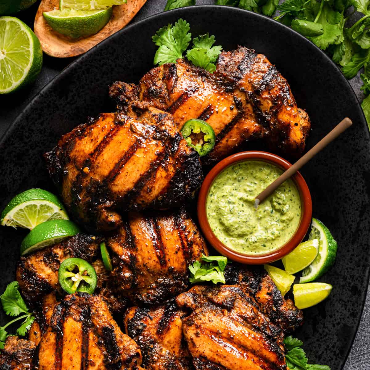 Grilled Peruvian chicken thighs with char marks on a black plate, garnished with lime wedges, cilantro, and sliced jalapeño. A small bowl of creamy green sauce sits on the plate beside the chicken.