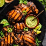 Grilled, seasoned Peruvian chicken thighs on a black platter, garnished with lime wedges, jalapeño slices, and fresh herbs. A bowl of green creamy sauce with a spoon sits beside the chicken.