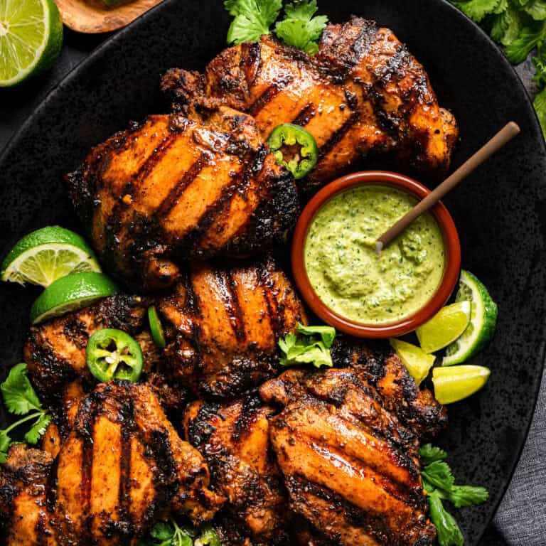 Grilled, seasoned Peruvian chicken thighs on a black platter, garnished with lime wedges, jalapeño slices, and fresh herbs. A bowl of green creamy sauce with a spoon sits beside the chicken.