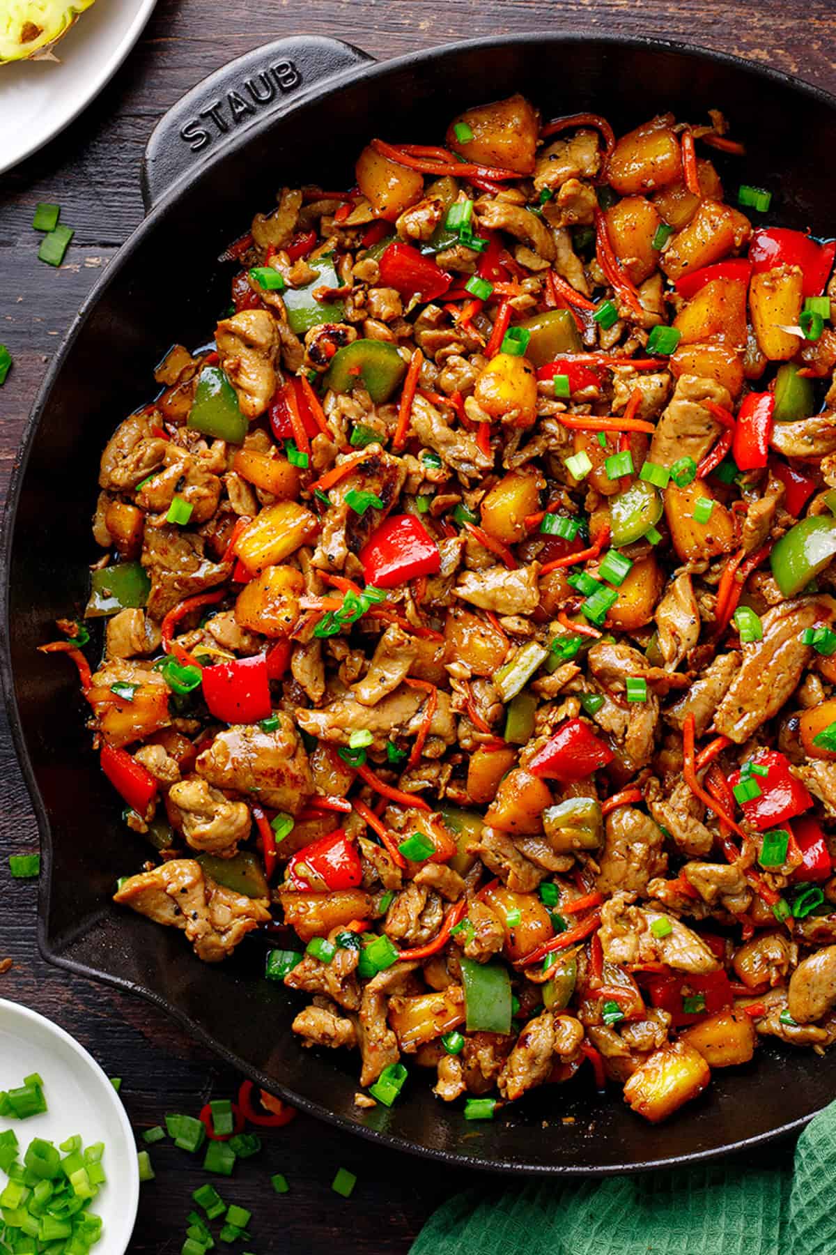 A skillet filled with stir-fried pineapple pork, bell peppers, carrots, chunks of pineapple, and green onions, garnished with chopped scallions; a colorful and appetizing dish on a dark wooden surface.