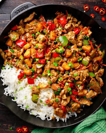 A cast iron skillet filled with stir-fried pineapple pork, bell peppers, and vegetables on one side, and white rice on the other, garnished with chopped herbs.