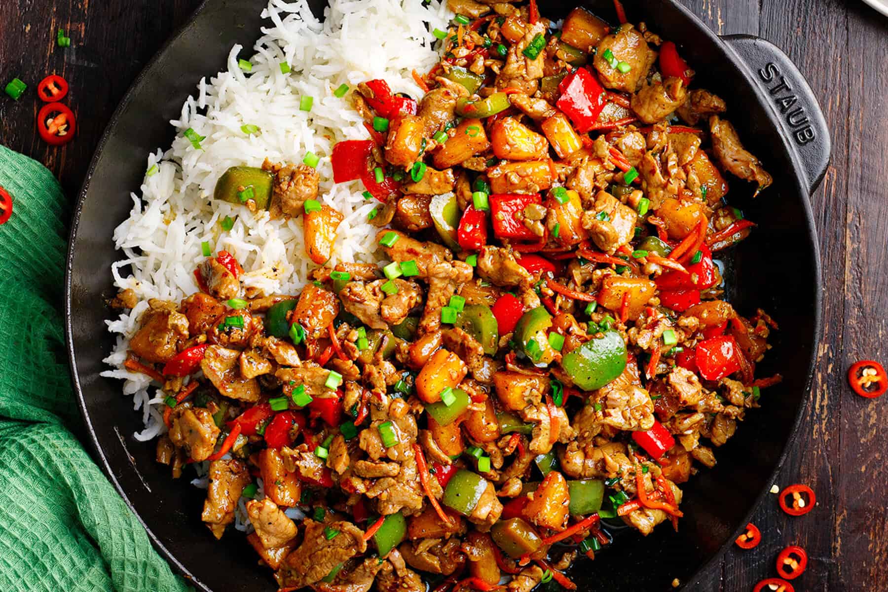 A skillet filled with stir-fried pineapple pork, colorful bell peppers, and vegetables next to a serving of white rice, all garnished with chopped green onions on a dark wooden table.