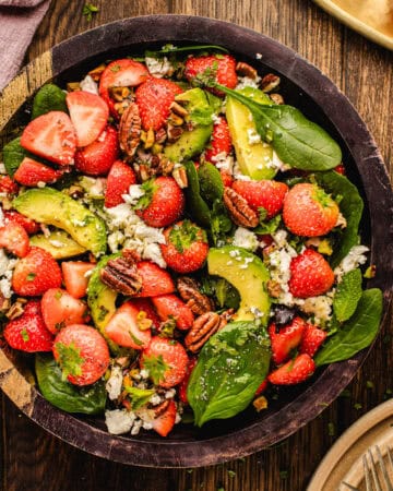 A wooden bowl filled with a fresh salad of spinach leaves, whole and halved strawberries, avocado slices, pecans, and crumbled feta cheese on a rustic wooden table.