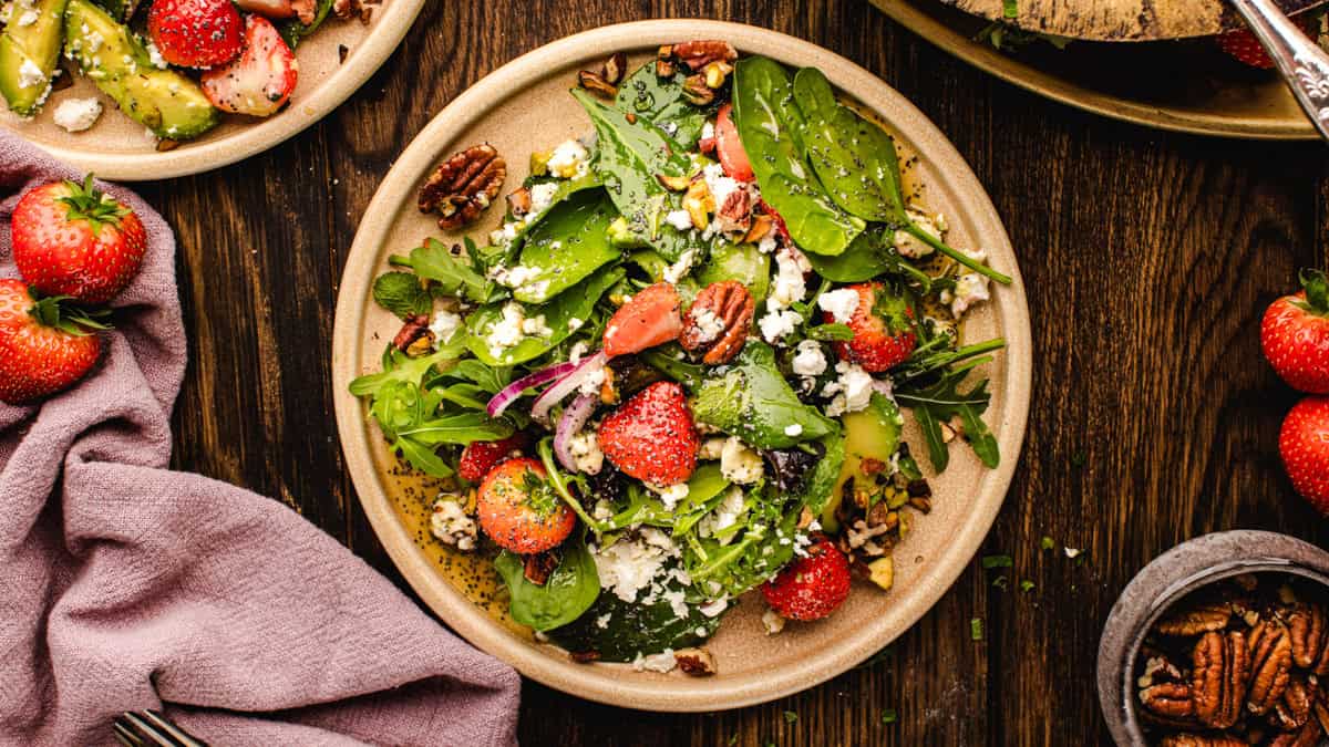 A fresh strawberry spinach salad on a beige plate features spinach, arugula, strawberries, pecans, avocado, and crumbled feta. Surrounding the plate are whole strawberries, a purple napkin, and a bowl of pecans on a wooden table.
