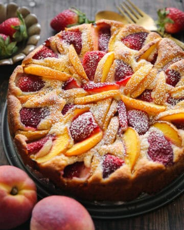 A round strawberry peach cake topped with sliced peaches and strawberries, dusted with powdered sugar, sits on a dark surface.