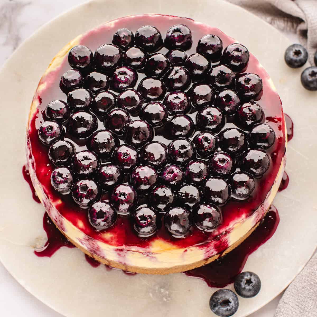 A round cheesecake topped with glossy blueberries and blueberry sauce, sitting on a light-colored plate. 