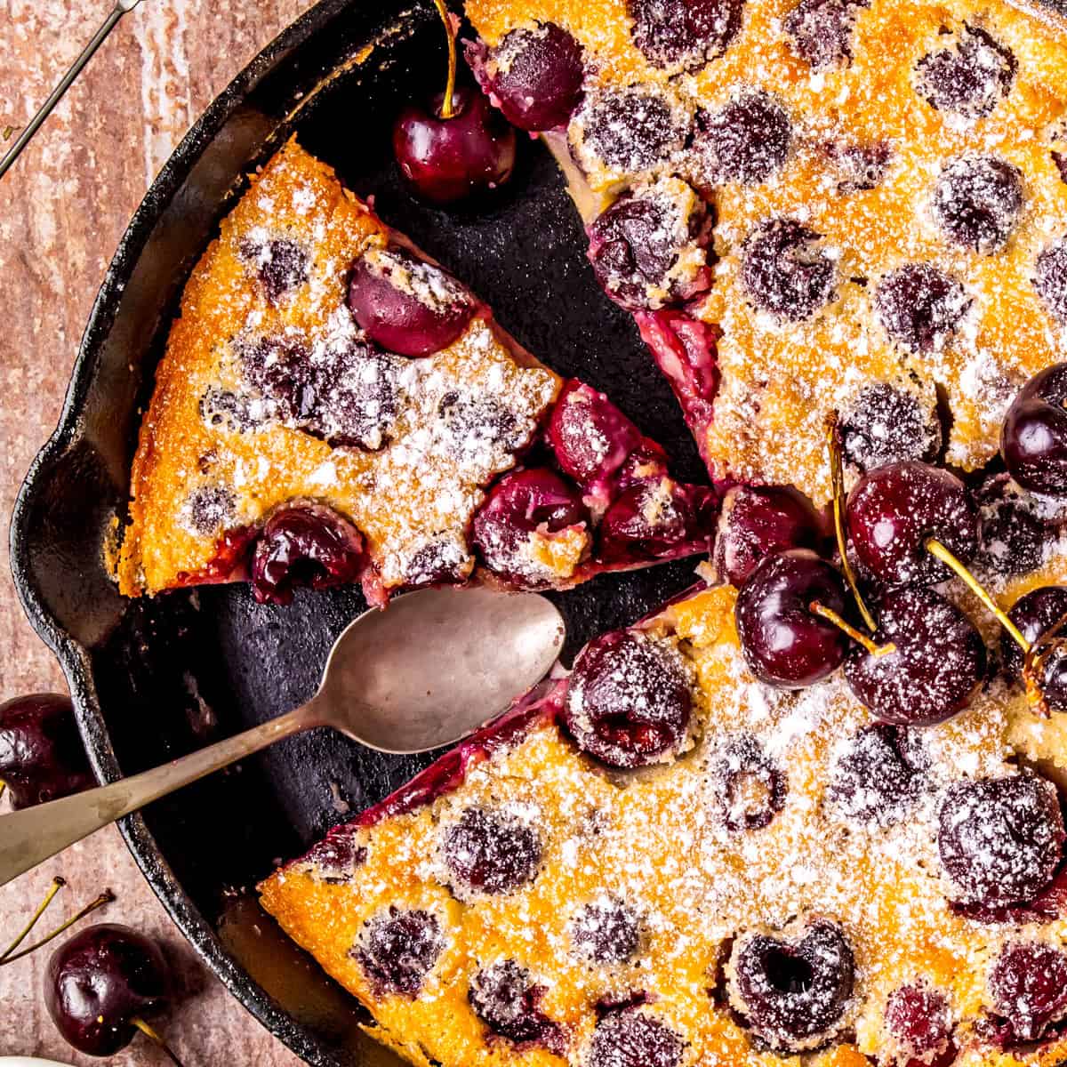A cast iron skillet holds a freshly baked cherry clafoutis, topped with fresh cherries and powdered sugar. A serving spoon rests in the pan, with a slice missing. 