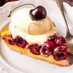A slice of cherry clafoutis topped with a scoop of vanilla ice cream and a whole cherry, served on a white plate with a silver spoon.