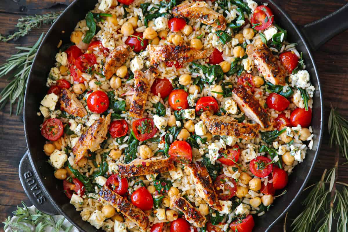 A skillet filled with a colorful mix of seared chicken strips, cherry tomatoes, chickpeas, spinach, feta cheese, and herbs on a wooden table.