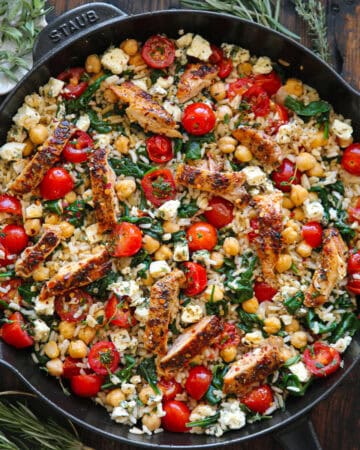 A skillet filled with seared chicken strips, chickpeas, cherry tomatoes, spinach, rice, and crumbled feta cheese, garnished with fresh herbs.