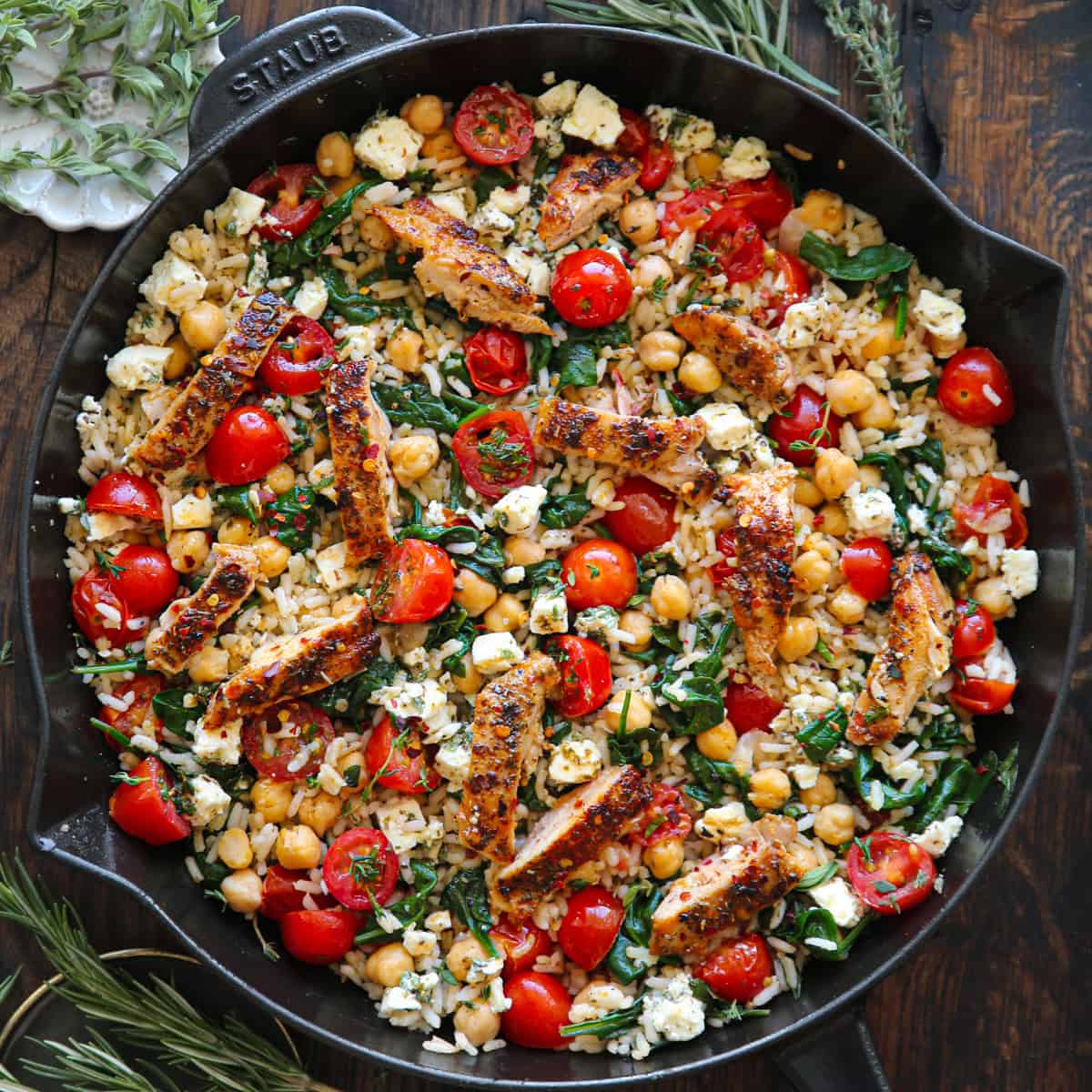 A skillet filled with seared chicken strips, chickpeas, cherry tomatoes, spinach, rice, and crumbled feta cheese, garnished with fresh herbs.