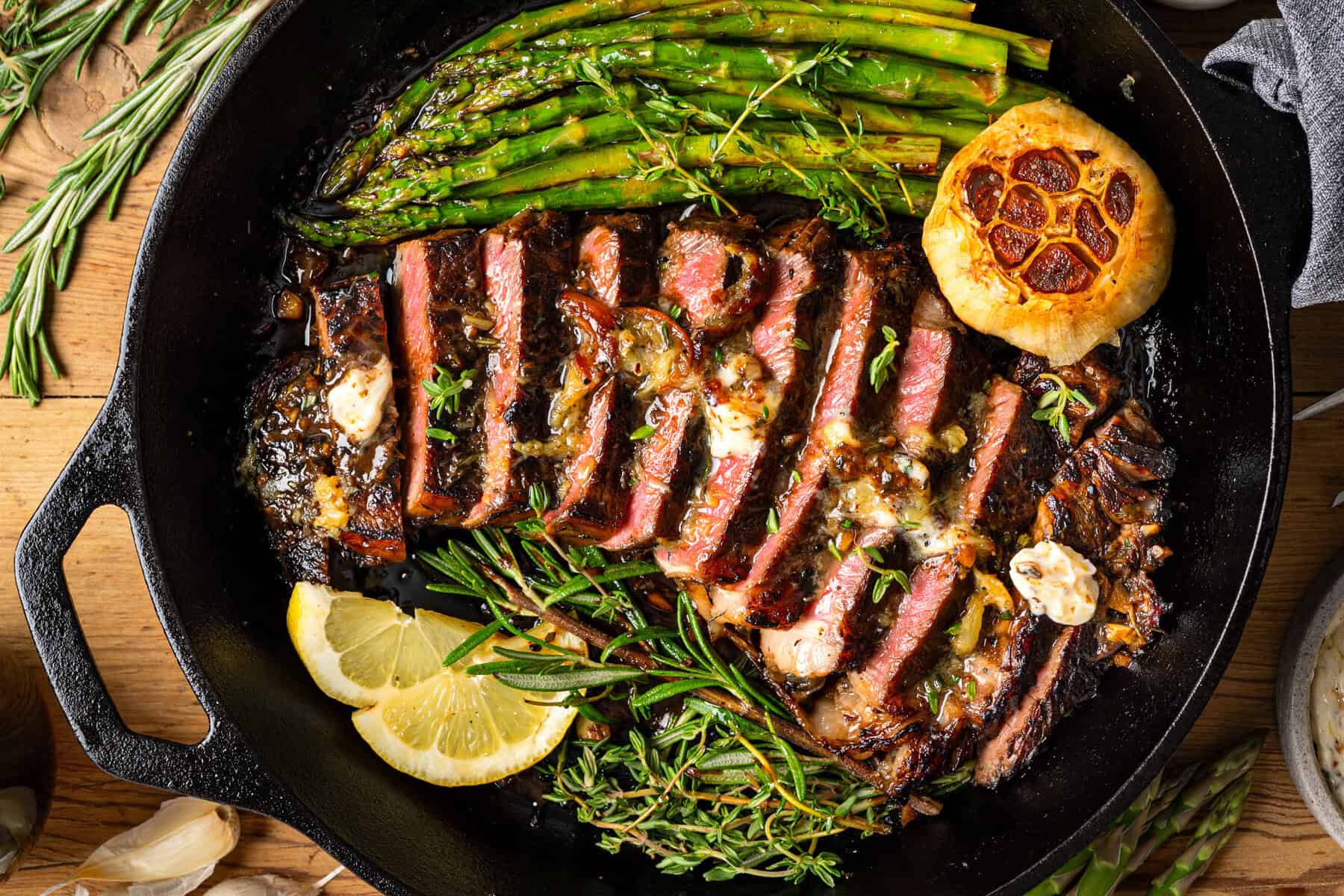 15 Steak Recipes Dad Will Love A cast iron skillet with sliced steak topped with herbs and butter, grilled asparagus, roasted garlic, fresh rosemary, and lemon slices on a wooden table.