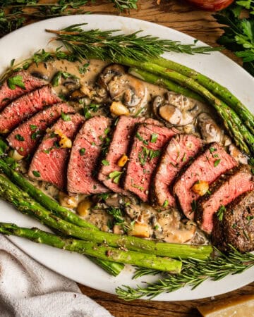 Sliced medium-rare steak topped with creamy mushroom sauce, garnished with herbs, and served with roasted asparagus on a white oval plate, surrounded by fresh herbs on a wooden surface.