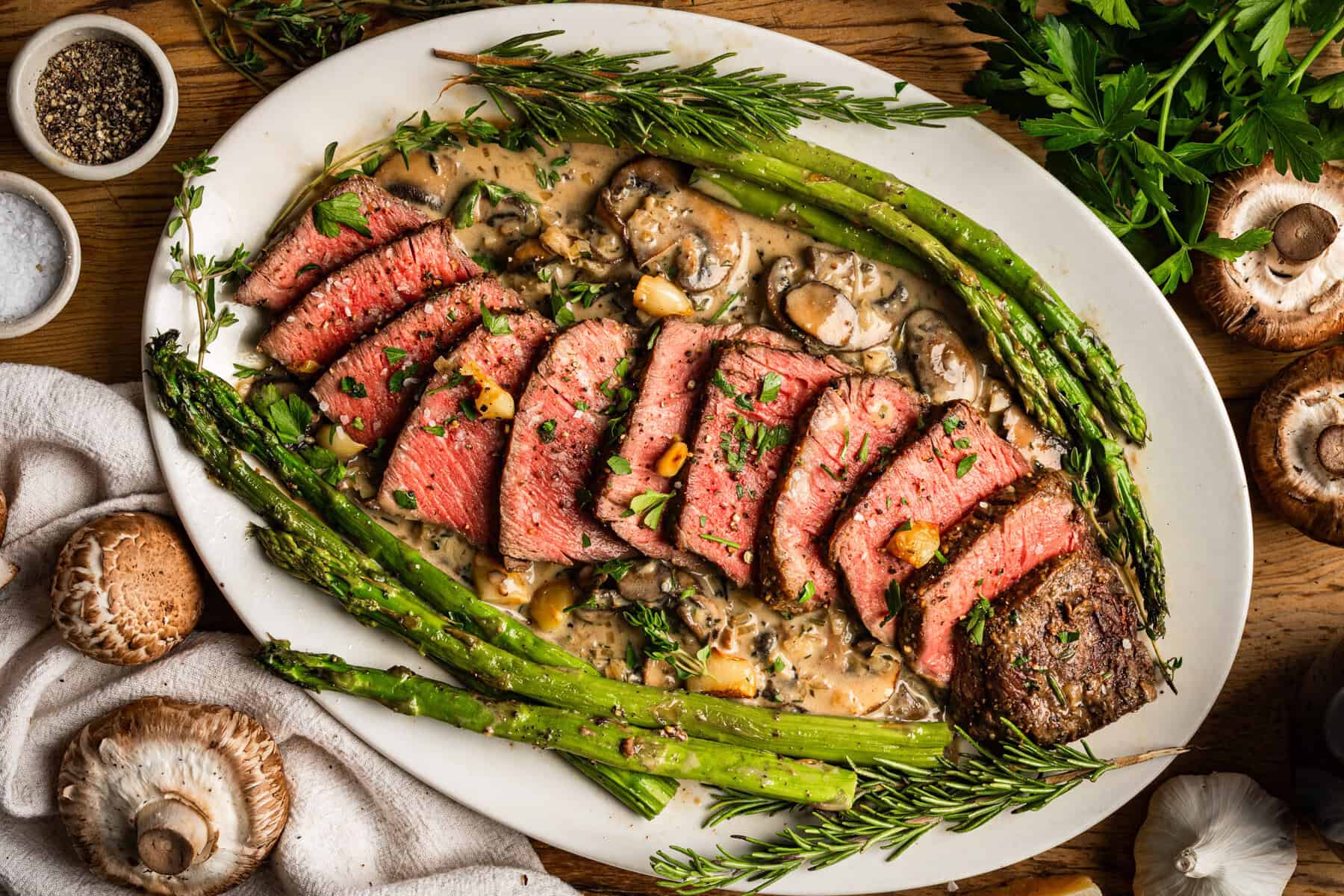 15 Steak Recipes Dad Will Love Sliced medium-rare steak served on a white oval plate with creamy mushroom sauce, roasted asparagus, and fresh herbs; surrounded by mushrooms, salt, pepper, and parsley on a wooden table.